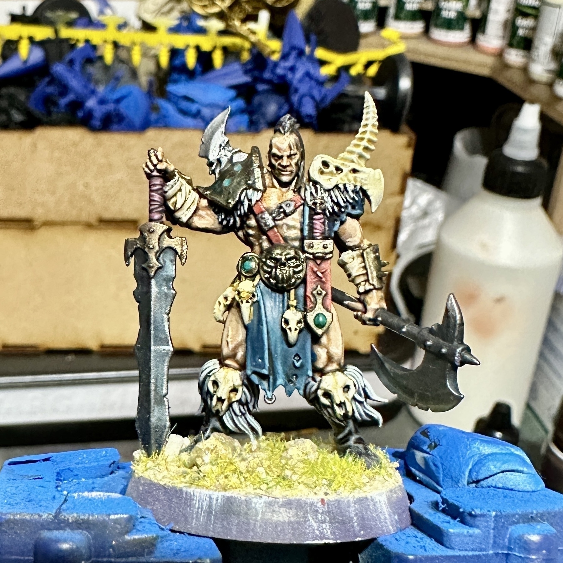 A mostly painted miniature fantasy warrior figure, armed with an axe and a sword, stands on a textured base amidst painting supplies, including brushes and paints, with other miniatures in the background.