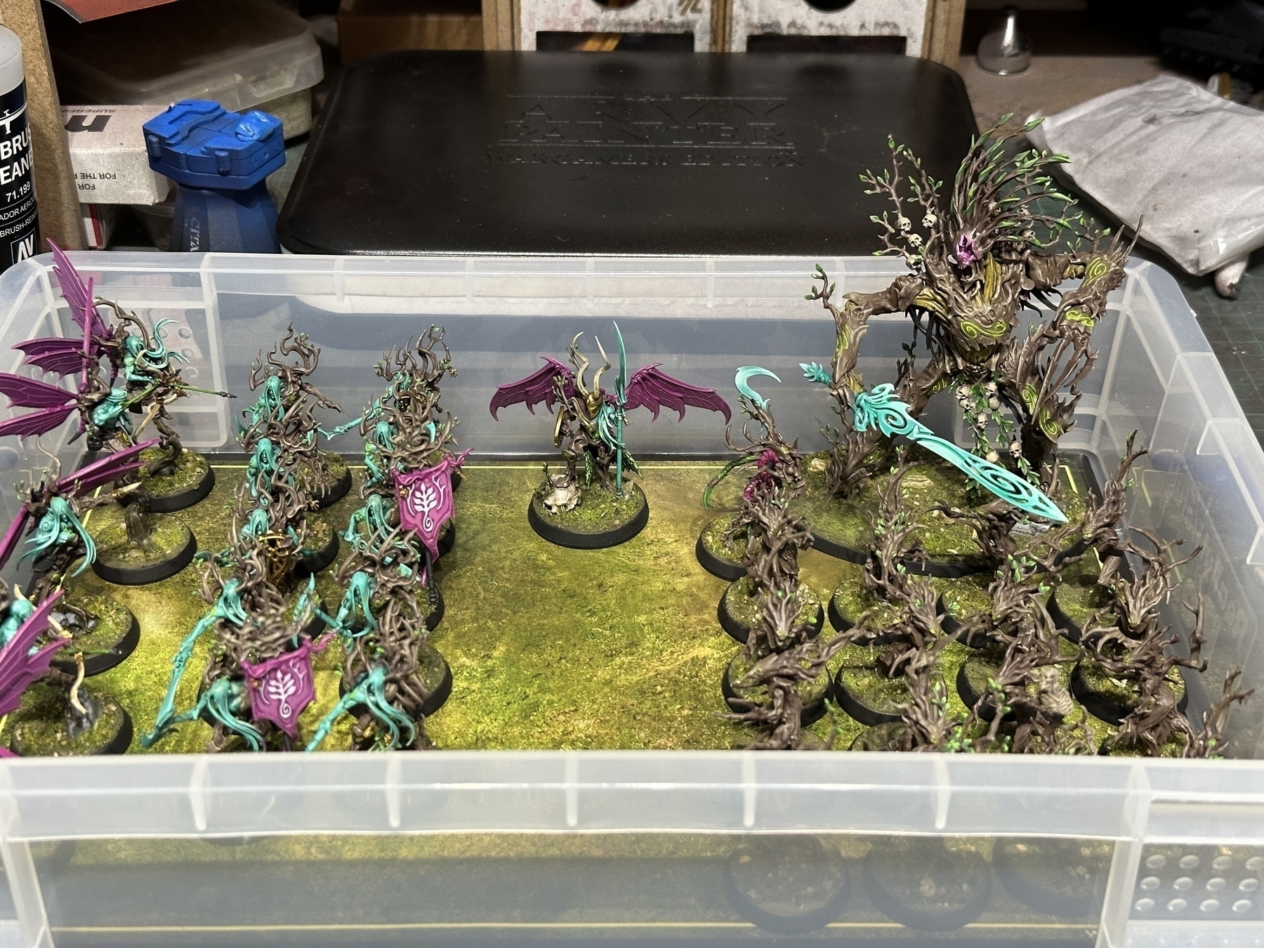 Auto-generated description: A collection of intricately painted miniature fantasy figures, featuring slender, elven-like characters with vibrant teal and purple detailing, stands on a surface mimicking a mossy ground,, is neatly organized in a plastic storage container on what appears to be a workbench cluttered with tools and other items.