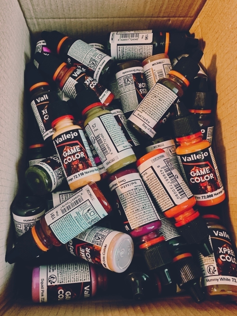 A cardboard box filled with a large selection of Vallejo miniature paints in dropper bottles, haphazardly piled on top of each other. There are a wide range of colours.