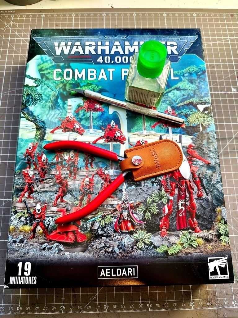 The Warhammer 40,000: Combat Patrol Aeldari box set of miniatures by Games Workshop, displayed on a cutting mat. On top of the box are clippers, a hobby knife, and Tamiya extra thin plastic cement